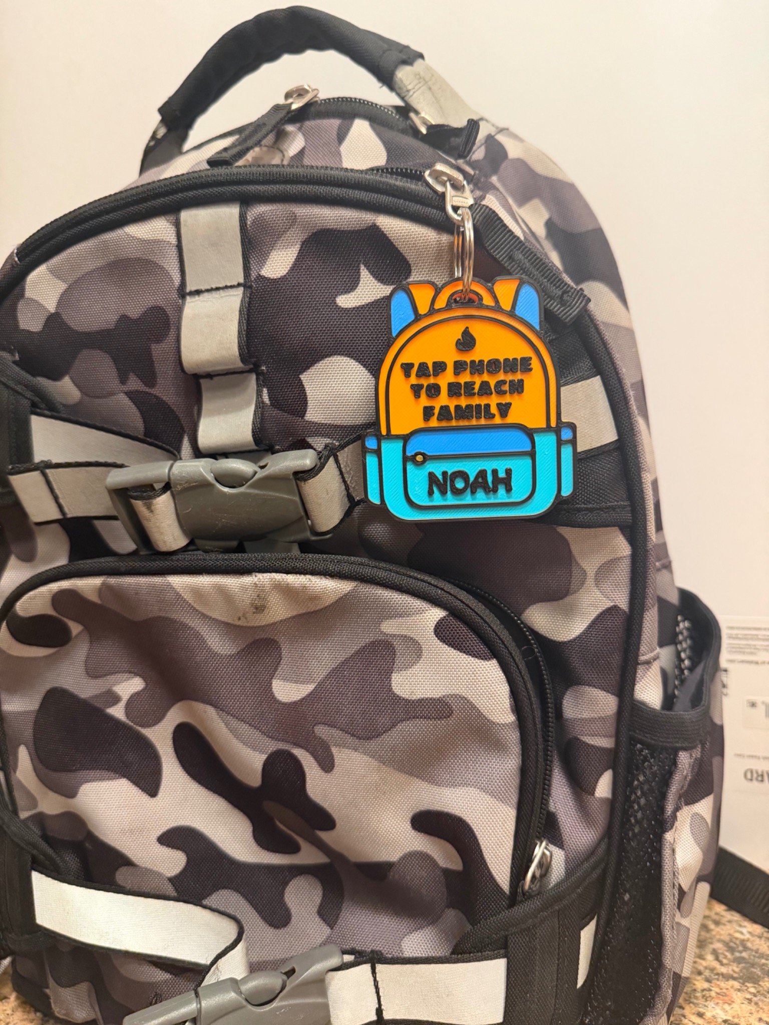 Real orange, blue and turquoise Fireside Prints NFC backpack tag personalized with NOAH, hanging on a camouflage backpack